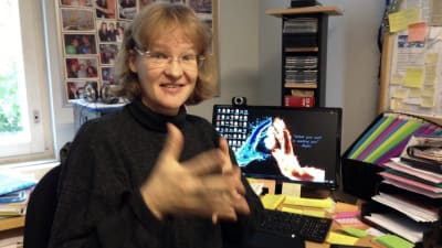 en kvinna sitter framför en dator och gör en rörelse med händerna på teckenspråk