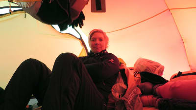 Kvinna med kort blont hår som sitter inne i ett tält. Det är fullt med kläder i tältet och i taket hänger också saker.