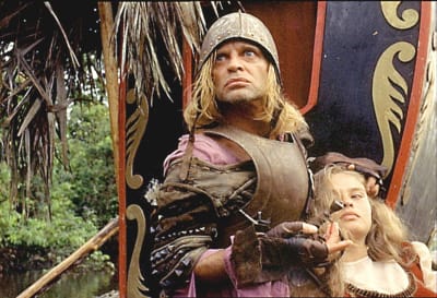 Haarnuskaan pukeutunut mies tuijottaa tuimasti ja pitelee kainalossaan vaaleaa naista, taustalla viidakkoa; kuva elokuvasta Aguirre.