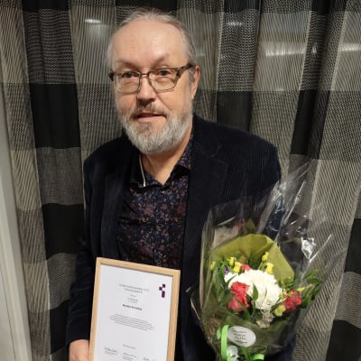 Mies pitelee käsissään kunniakirjaa ja kukkakimppua.