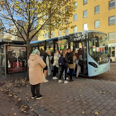 Ihmisiä menossa bussipysäkiltä sisään vaaleansiniseen bussiin. Etualalla henkilö, jolla on päässään turkoosi huivi ja yllään vaaleanruskea takki. Taustalla mukulakivikatu ja kerrostalon seinät, joista toisessa lukee optikko. Bussipysäkin vieressä kasvaa puu, jonka keltaisia lehtiä on pudonnut maahan.