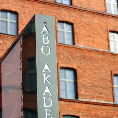 i förgrunden en skylt med texten Åbo Akademi. I Bakgrunden en röd tegelbyggnad.