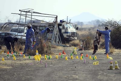 Några personer i blå overaller utför undersökningar efter massakern i Marikana. Gula, gröna och orange markeringar på marken. Några fordon i bakgrunden. 