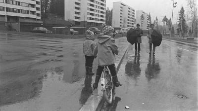 År 1970 i en förort i Helsingfors. Höghus. Barn på cykel och två män med paraply på en regnig gata.