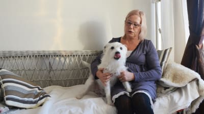 En kvinna sitter i en soffa och håller om en liten vit hund. Kvinnan tittar på hunden som slickar sig om munnen.
