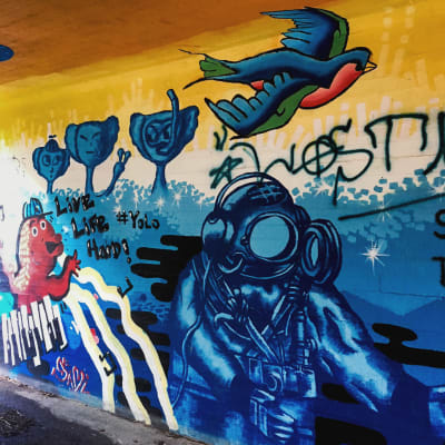 Värikäs seinämaalaus alikulkutunnelissa, jossa tekstit live life hard, yolo ja kuva astronautista ja linnusta.