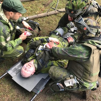 Sårad ligger på bår med tre rekryter som försöker hjälpa, under en militärövning.