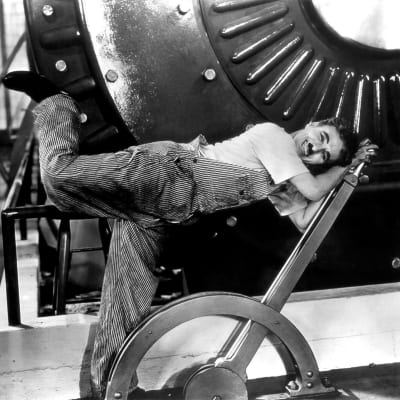 Työhaalariin pukeutunut mies (Charlie Chaplin) nojaa nauraen suureen säätövipuun, taustalla suuri kone. Kuva elokuvasta Nykyaika.