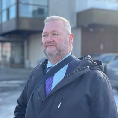 Mies katsoo kaukaisuuteen, taustalla rakennus ja autoja.