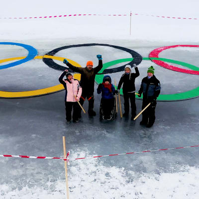 Joukko ihmisiä seisoo isojen jäähän tehtyjen ja värikkäiden olympiarenkaiden edessä. Osa vilkuttaa ylöspäin kameralle.