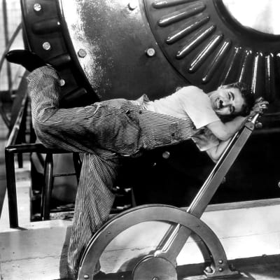 Työhaalariin pukeutunut mies (Charlie Chaplin) nojaa nauraen suureen säätövipuun, taustalla suuri kone. Kuva elokuvasta Nykyaika.