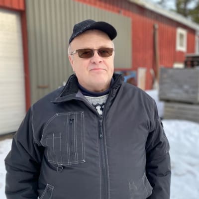 Man med keps och svart jacka framför ett uthus. 