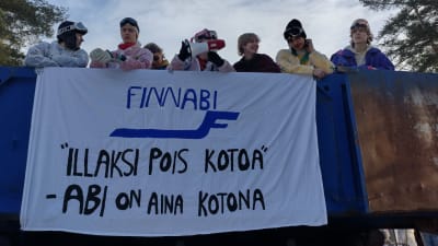Ungdomar på lastbilsflak i Virkby, de är klädda i skiddress för utförsåkning . På affisch står det på finska (översatt) FinnAbi, hemifrån på kvällen, abirna är alltid hemma.