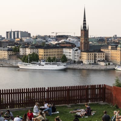 Människor sitter på gräsmattan i Ivar Los park i Stockholm och blickar ut mot stadens centrum en kväll i juli 2021.