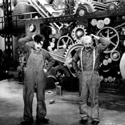 Kaksi työhaalareihin pukeutunutta miestä raapii päätään mietteliäinä valtavan koneen edessä. Kuva elokuvasta Nykyaika.