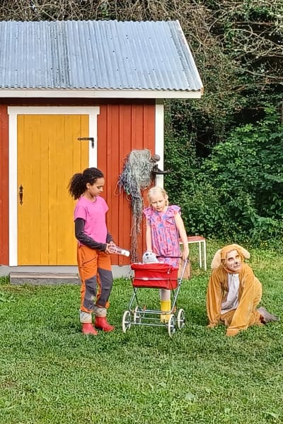 Två flickor med en barnvagn och en person utklädd till en hund står framför en röd liten stuga.