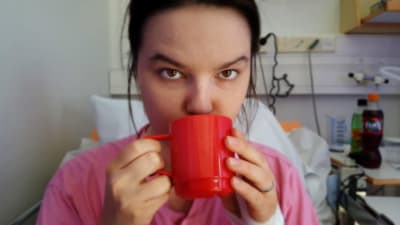 Pernilla Bergman sitter i sin sjukhussäng, ser in i kameran och dricker ur en röd mugg