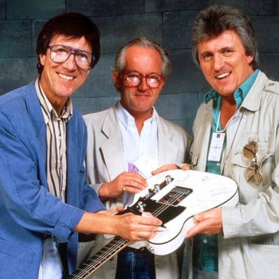 Hank Marvin, Brian Bennett och Bruce Welch i The Shadows håller i en elgitarr som de signerar.