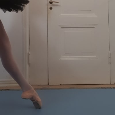 Nainen seisoo balettihameessa ja balettitossuissa oven edessä. 
