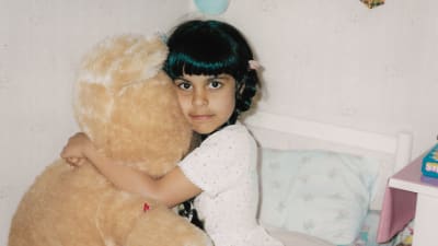 Ett barn med långt svart hår sitter på en säng och kramar en stor ljusbrun nalle.