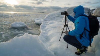 En person i vinterrock fotograferar vid havet, is och snö, sol.