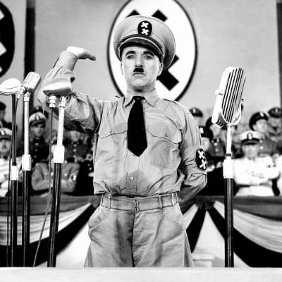Univormupukuinen, hitlerviiksinen mies (Charlie Chaplin) seisoo puhujanpaikalla mikrofonien edessä ja tekee eräänlaista Hitler-tervehdystä. Kuva elokuvasta Diktaattori.