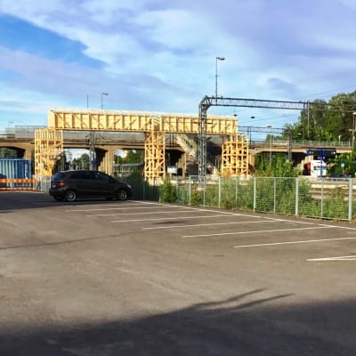 I förgrunden en tom asfalterad parkering. Längre borta syns en bro över en bangård samt en tillfälllig träbro. Karis järnvägsstations, en gul stockbyggnad, syns till höger.