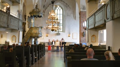 Längst fram syns altaret i Borgå domkyrka och fyra sjungande kvinnor. I bänkraderna sitter en hel del människor, främst unga par.