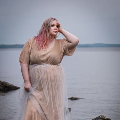 Kvinna i beige klänning och blont och rosa hår. Hon står på klippor framför havet och tittar ut till höger och håller handen mot pannan och sidan av huvudet.