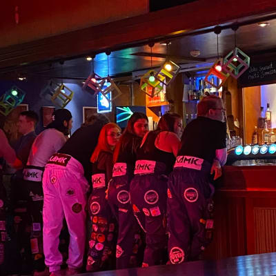 Rivi opiskelijoita nojaa baaritiskiin. Kaksi heistä katsoo taakseen kohti kameraa. 
