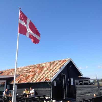 Danska flaggan vajar utanför lantligt hus, blå himmel.