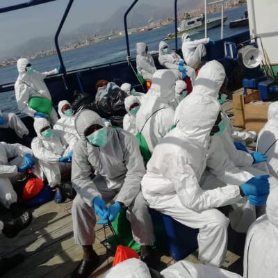 De migranter som räddats av det spanska räddningsfartyget Aita Mari, fick klä på sig skyddsdräkter och fördes vidare till den italienska passagerarfärjan på söndagen. 