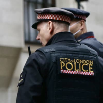 Poliser i London står med ryggen mot kameran. På deras rygg står "City of London police". De är klädda i uniform.