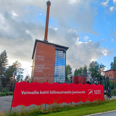 Taustalla punatiilisiä tehdasrakennuksia. Etualalla verkkoaitaa, jossa suuri kyltti "Voimalla kohti hiilineutraalia Joensuuta, Savon Voima". Puolipilvinen taivas.
