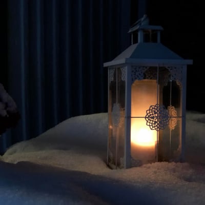 Kynttilä palaa lyhdyssä, joka on lumen peittämällä pöydällä.