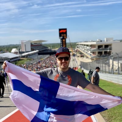 En glad man med solglasögon och keps och finländsk flagga dtår på en sportplan.