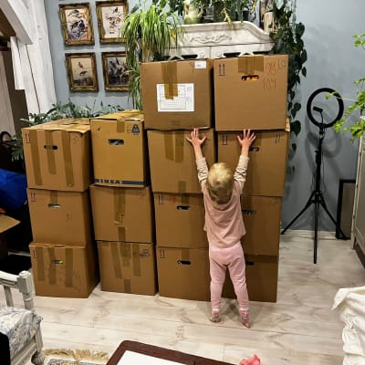 Ett barn på tv-tre år står med ryggen mot kameran och sträcker sig på tå för att försöka nå det stora "berget" av bruna papplådor. I lådorna finns kläder till barn i Ukraina.