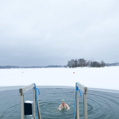 Vappu Hietala, kassör för Seurasaaren uimarit ry, badar i isvaken på Fölisöns utebad. Hon bär en färggrann mössa.