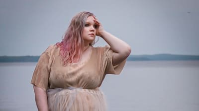 Kvinna i beige klänning och blont och rosa hår. Hon står på klippor framför havet och tittar ut till höger och håller handen mot pannan och sidan av huvudet.