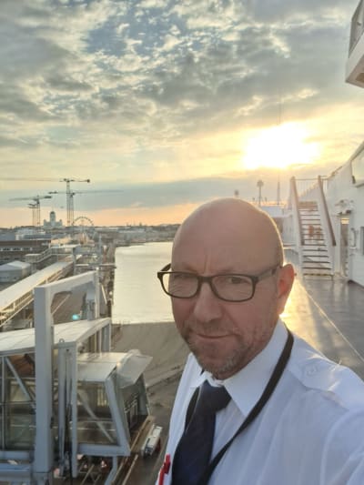 En man utan hår, med gråsvart kort skägg och mustasch, glasögon, vit skjorta och slips tittar in i kameran. Han står på ett fartygsdäck med solen och siluetten av Helsingfors i bakgrunden. 