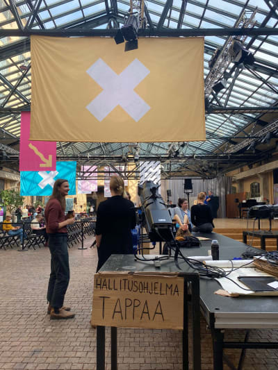 Teaterhögskolans entré. I förgrunden en skylt där det står "hallitusohjelma tappaa", regeringsprogrammet dödar, och i bakgrunden studerande som står och pratar.