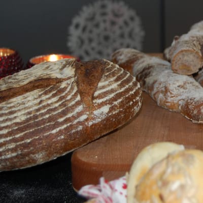 Ett skärbräde med hembakt bröd i olika utformning, några ljusa semlor med frön på, några grovt tvinnade baguetter och limpor som snittats för vackra mönster.