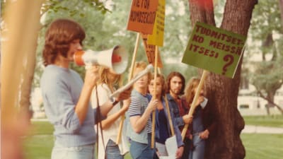 Nuoria aikuisia 1974 mielenosoituskylttien ja megafonin kanssa seisomassa puistossa.