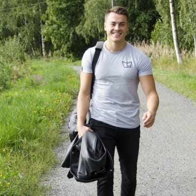 En man iklädd svarta jeans och en grå t-skjorta går på en grusväg kantad med grönt gräs och blommor. Han ler och ser glad ut.