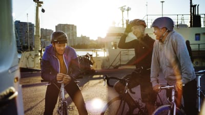 Tre män står vid sina cyklar i hamnen, solen skiner i bakgrunden.