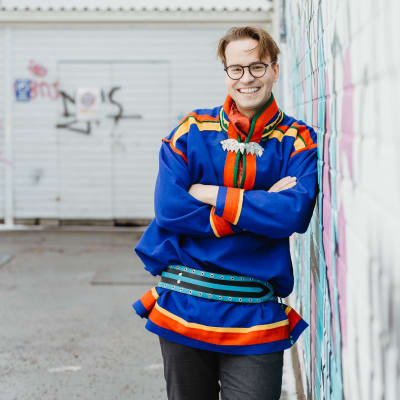 Janne Hirvasvuopio är klädd i en samekolt och ser glad ut. Han lutar ena axeln mot en trävägg med klotter på.