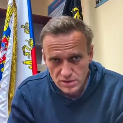 Aleksei Navalnyi katsoo vakavana kameraan. Hänellä on yllään sininen pusero, Taustalla huoneessa näkyy lippuja.ja ilmoitustaulu.