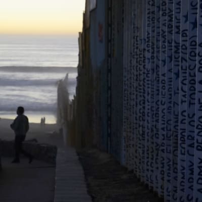 Muren som delar Mexico och USA. Ansikten och namn står inskrivna på muren. I bakgrunden syns en sandstrand och havet.