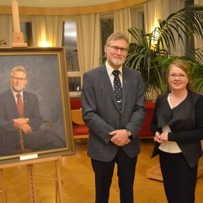 Ett porträtt av pensionerade stadsdirektören i Hangö Jouko Mäkinen. Bredvid tavlan står Mäkinen och konstnären Arja Järvilehto. I bakgrunden en stor grönväxt samt en julgran.