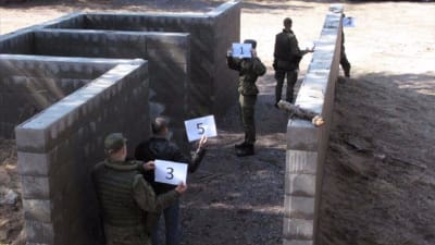 Några män i militärkläder står invid väggar uppmurade av betongblock, håller nummerskyltar i händerna.